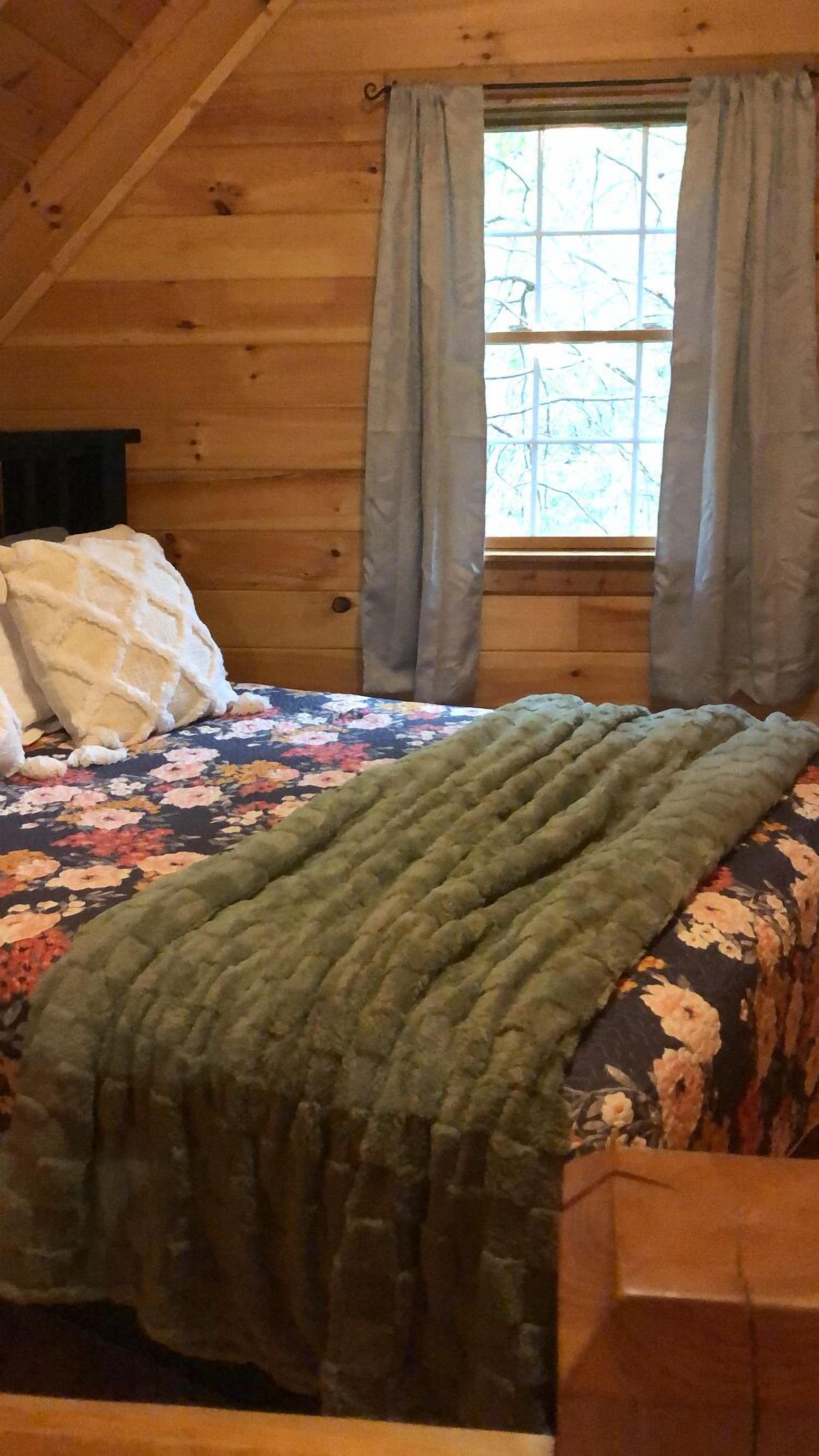 Une pièce chaleureuse avec un plafond en bois, un lit recouvert d’une courtepointe à motifs floraux et d’un plaid vert, et une fenêtre avec des rideaux.