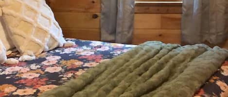 Une pièce chaleureuse avec un plafond en bois, un lit recouvert d’une courtepointe à motifs floraux et d’un plaid vert, et une fenêtre avec des rideaux.