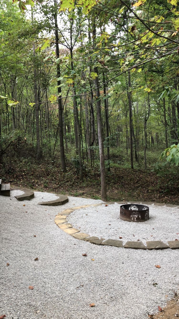 A forested area with a gravel path, a circular stone bench, and a metal fire pit.