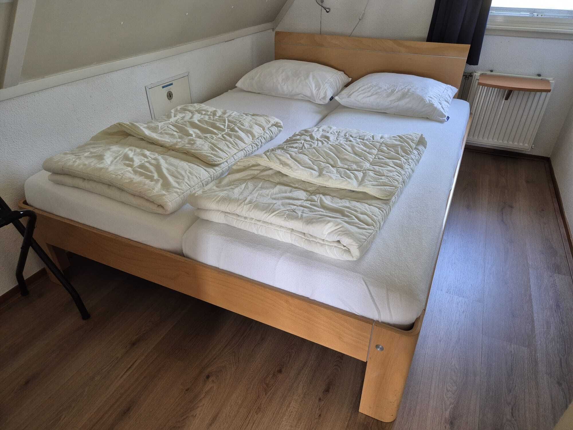 Uma cama de solteiro com estrutura de madeira e roupa de cama branca, um radiador e uma janela com cortinas.