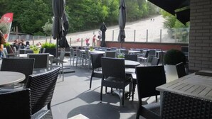 An outdoor patio area with round tables and chairs, umbrellas, and a view of a beach and trees.