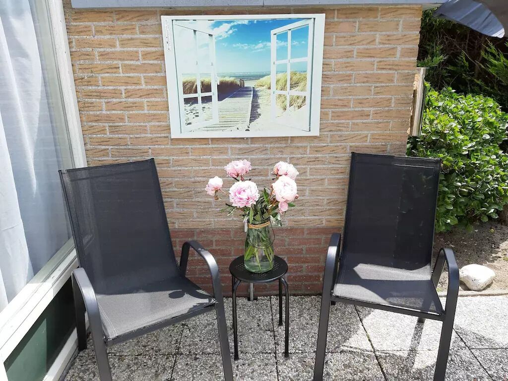 Dos sillas negras, una mesita con un florero de flores rosas y un cuadro enmarco con una playa en una pared de ladrillo.