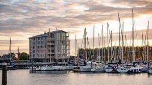 Un port de plaisance où sont amarrés de nombreux voiliers, un bâtiment moderne doté de balcons et un ciel au coucher du soleil.