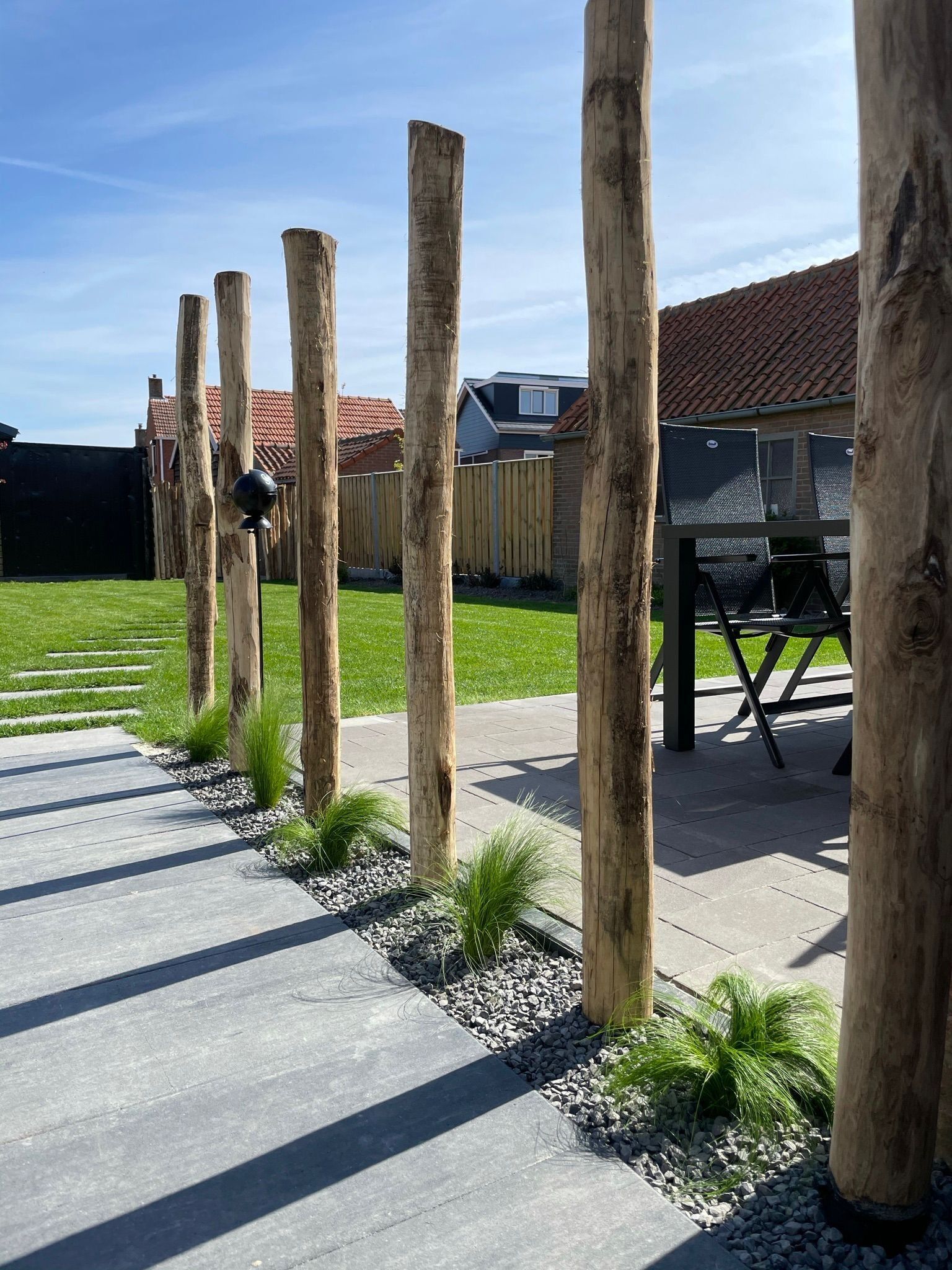 A garden with wooden posts, a concrete pathway, and a patio area with chairs.