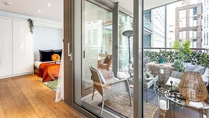 Un interior moderno con un vidrio que da a un balcón exterior. En el interior, hay una cama con una colcha roja, una silla de mimbre y una mesita con una lámpara. El balcón tiene una silla de mimbre, una mesa y plantas en macetas.