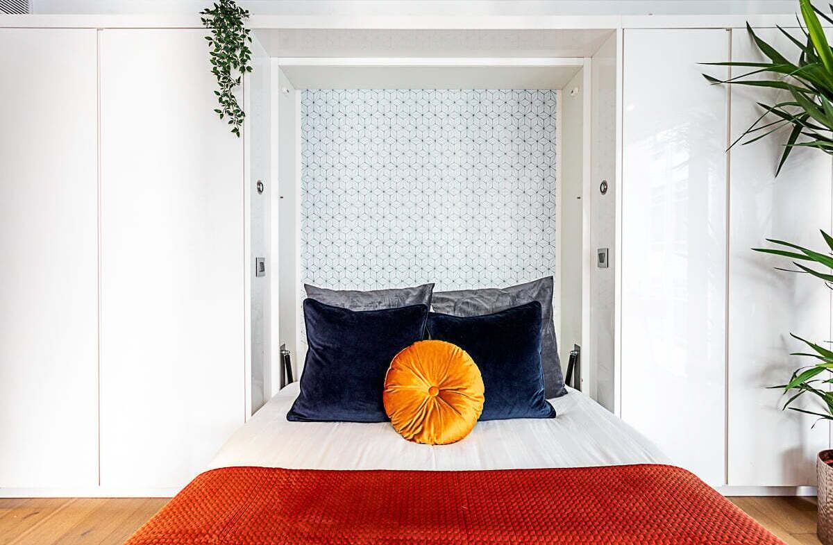 Une chambre à coucher avec un lit placé au centre d’un encadrement de porte, des murs blancs, un rideau à motifs géométriques et un tapis rouge.