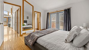 Une chambre avec un grand lit, une vue sur l’extérieur par une fenêtre et un couloir menant à une autre pièce.