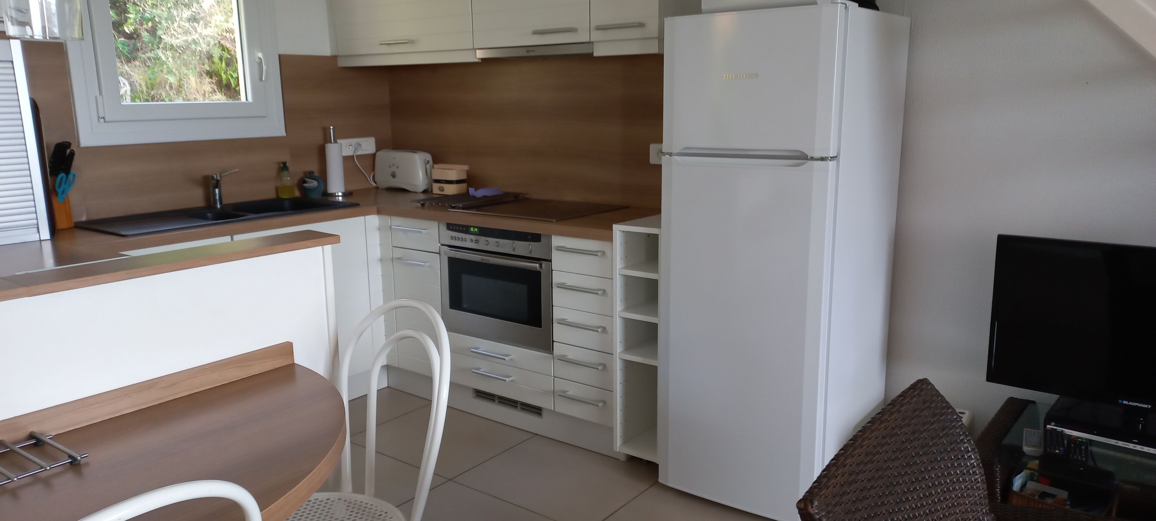 Een compacte keuken met witte kasten, een ingebouwde oven, een koelkast en een eethoek met een stoel.