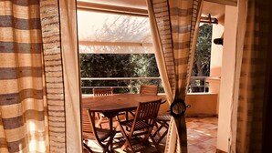 A balcony with a wooden table and chairs, a view of trees, and a patterned curtain. - Charming*** villa with heated pool, close to town center (Porto-Vecchio)