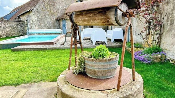 A rustic outdoor setting with a wooden barrel structure, a stone well, and a swimming pool in the background. - Gites-domainedupin, "gîte de la Closerie (Mosnes)