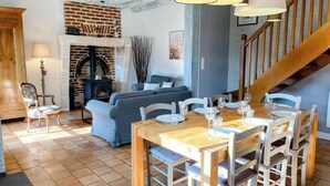 A cozy living room with a fireplace, a dining area with a wooden table and chairs, and a staircase leading to an upper floor. - Gites-domainedupin, "gîte de la Closerie (Mosnes)