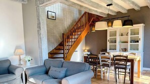 A living room with a grey sofa, wooden staircase, dining table with chairs, and a kitchen area with cabinets. - Gites-domainedupin, "gîte de la Closerie (Mosnes)