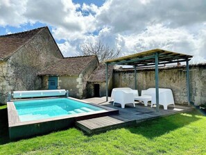 A swimming pool with a wooden deck, white outdoor furniture, and a stone building in the background.