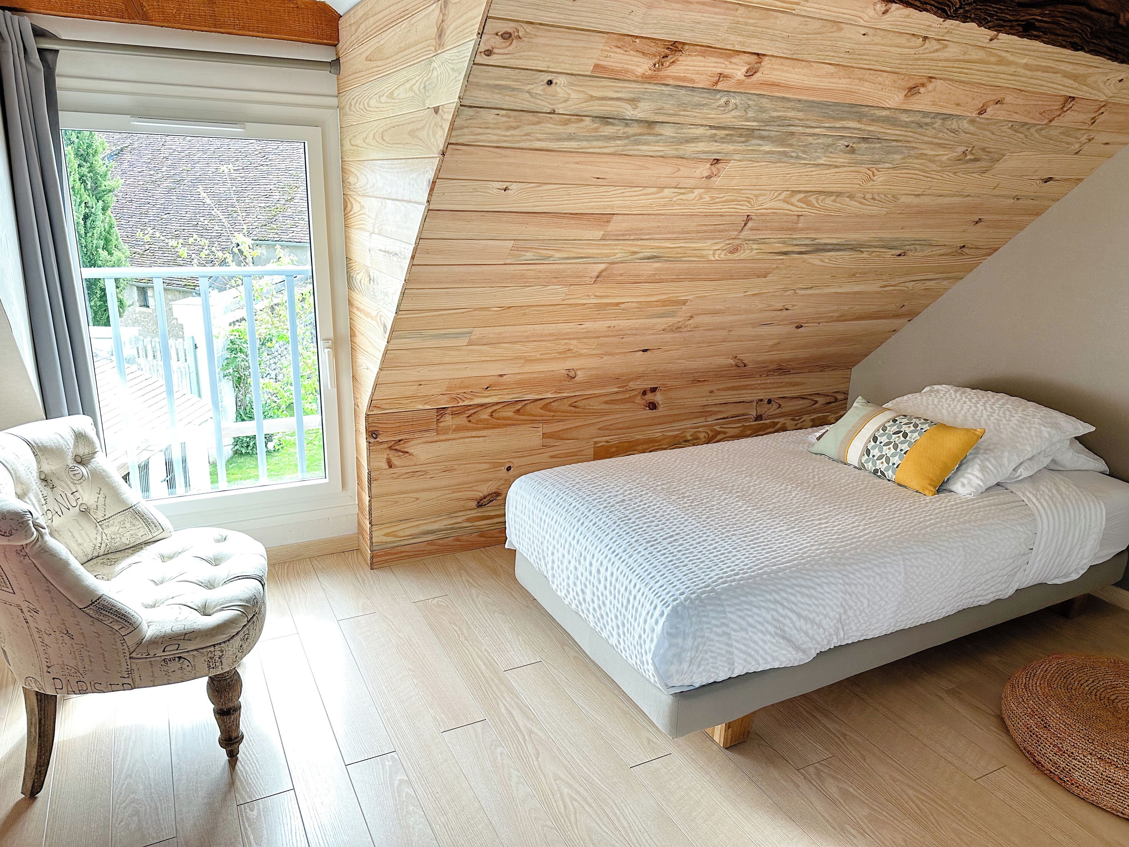 A bedroom with a sloped ceiling, a bed with white bedding, a chair with a patterned cushion, and a view of greenery outside.
