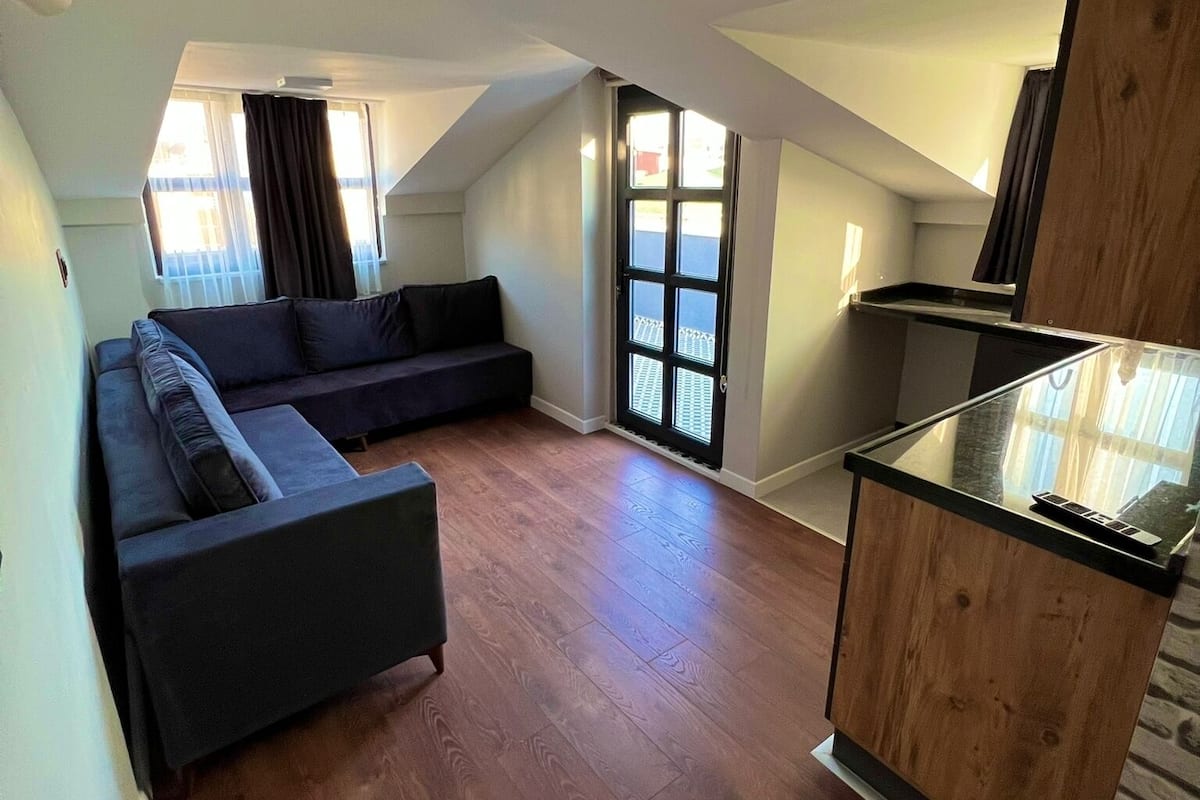 A spacious loft apartment with a large dark-colored sectional sofa, a wooden dining area, and a sliding glass door leading to a balcony.