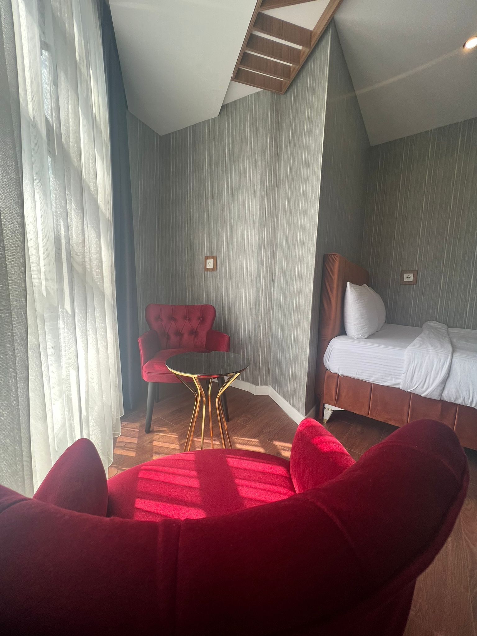 A hotel room with a bed, a red armchair, a small round table, and a window with sheer curtains.