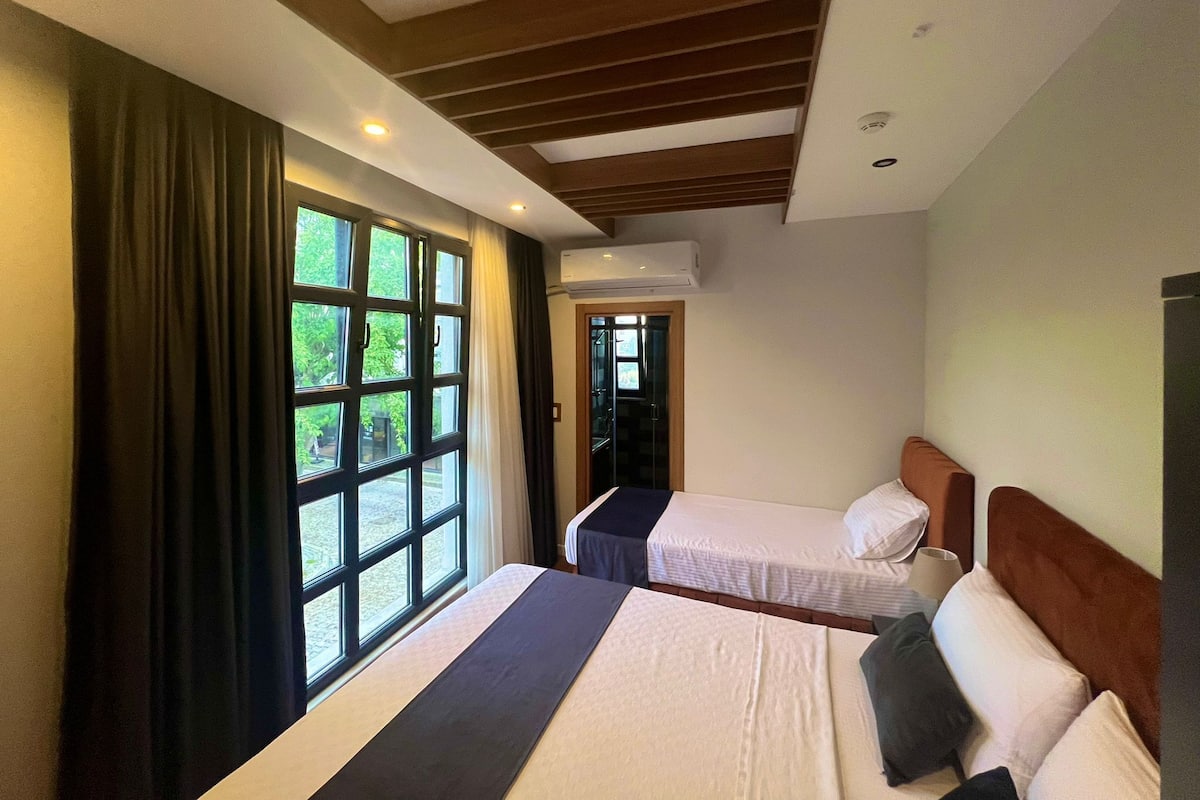 A hotel room with two beds, a large window with curtains, an air conditioning unit, and a wooden ceiling.