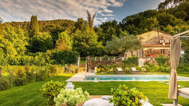 A poolside area with a stone house, lush greenery, and a wooden deck. - Villa Ester in Capannori (Capannori)