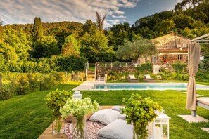 A poolside area with a stone house, lush greenery, and a wooden deck. - Villa Ester in Capannori (Capannori)
