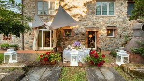 A stone building with a patio area featuring a table set for dining, surrounded by potted plants and outdoor furniture. - Villa Ester in Capannori (Capannori)