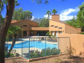 Zona de piscina con piscina cercada, sillas de descanso y un edificio de techo plano y paredes color beige.