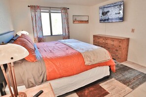 A bedroom with a bed, a wooden dresser, a lamp, a window with curtains, and a painting on the wall.