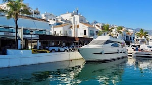 Una marina con uno yacht bianco ormeggiato, circondata da edifici bianchi con negozi e ristoranti, e palme.