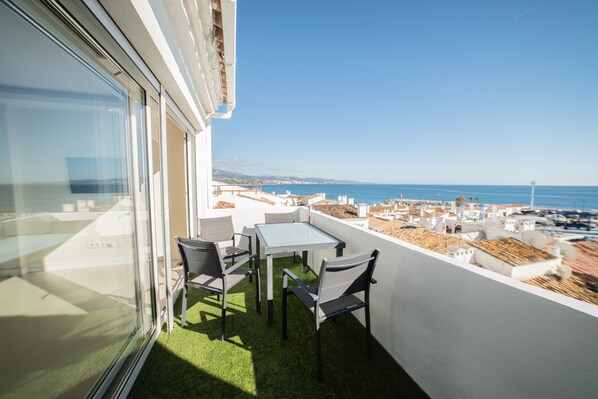 A balcony with a glass door, a small table, and two chairs. The balcony overlooks a coastal area with buildings and a clear sky.