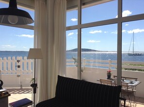 Une pièce dotée d’une grande fenêtre donnant sur la mer et sur un voilier. On y trouve un lit avec un oreiller à rayures noires, une table avec des chaises et une lampe.