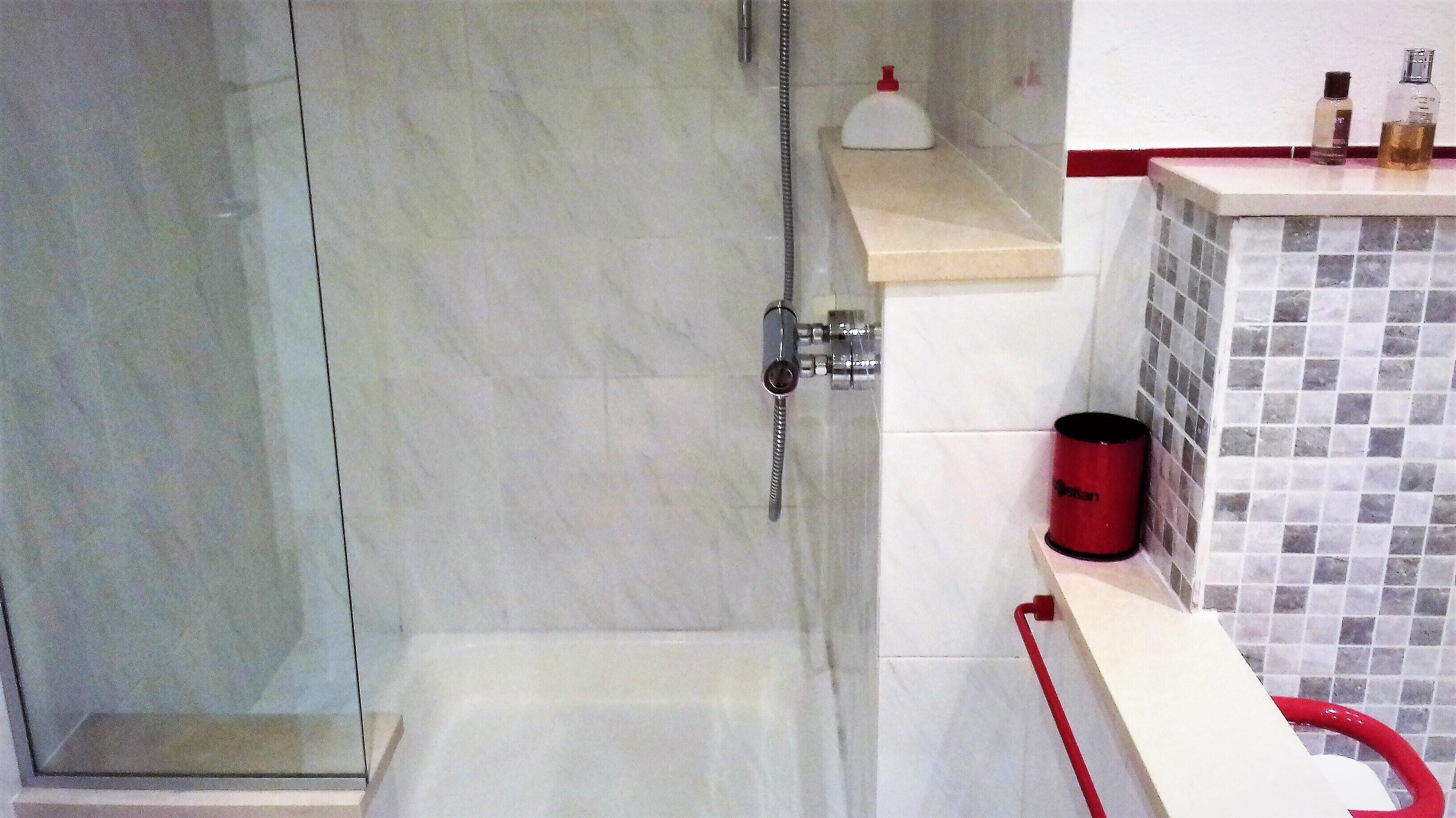 A bathroom with a glass shower door, a tiled wall, and a red bucket on a shelf.