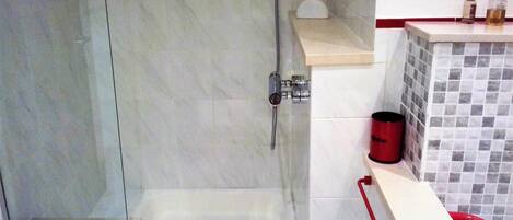 A bathroom with a glass shower door, a tiled wall, and a red bucket on a shelf.