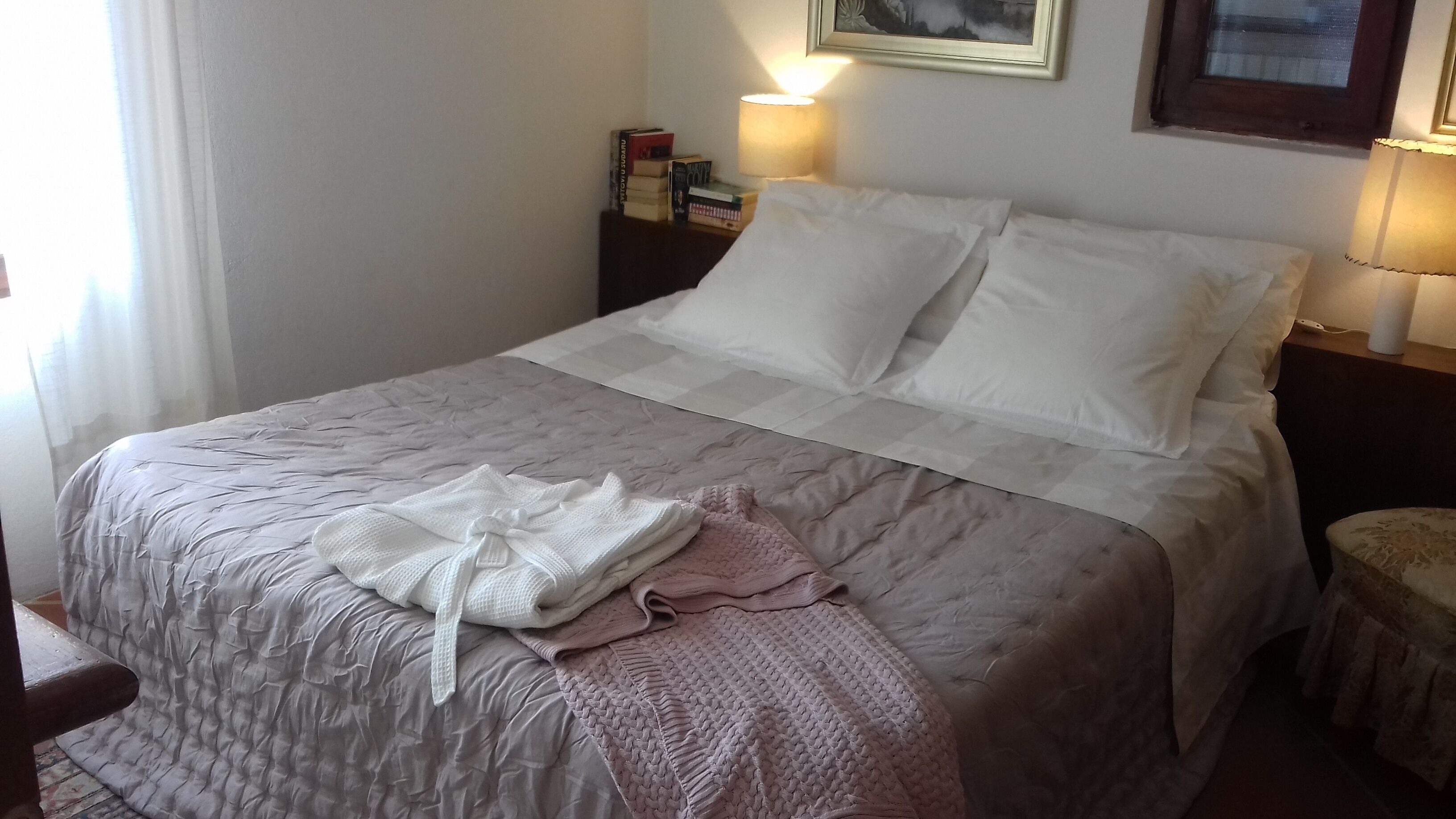 Ein ordentlich bezogenes Bett mit einer weiß-rosa Decke, ein Nachttisch mit Büchern und einer Lampe sowie ein Fenster mit Vorhängen.