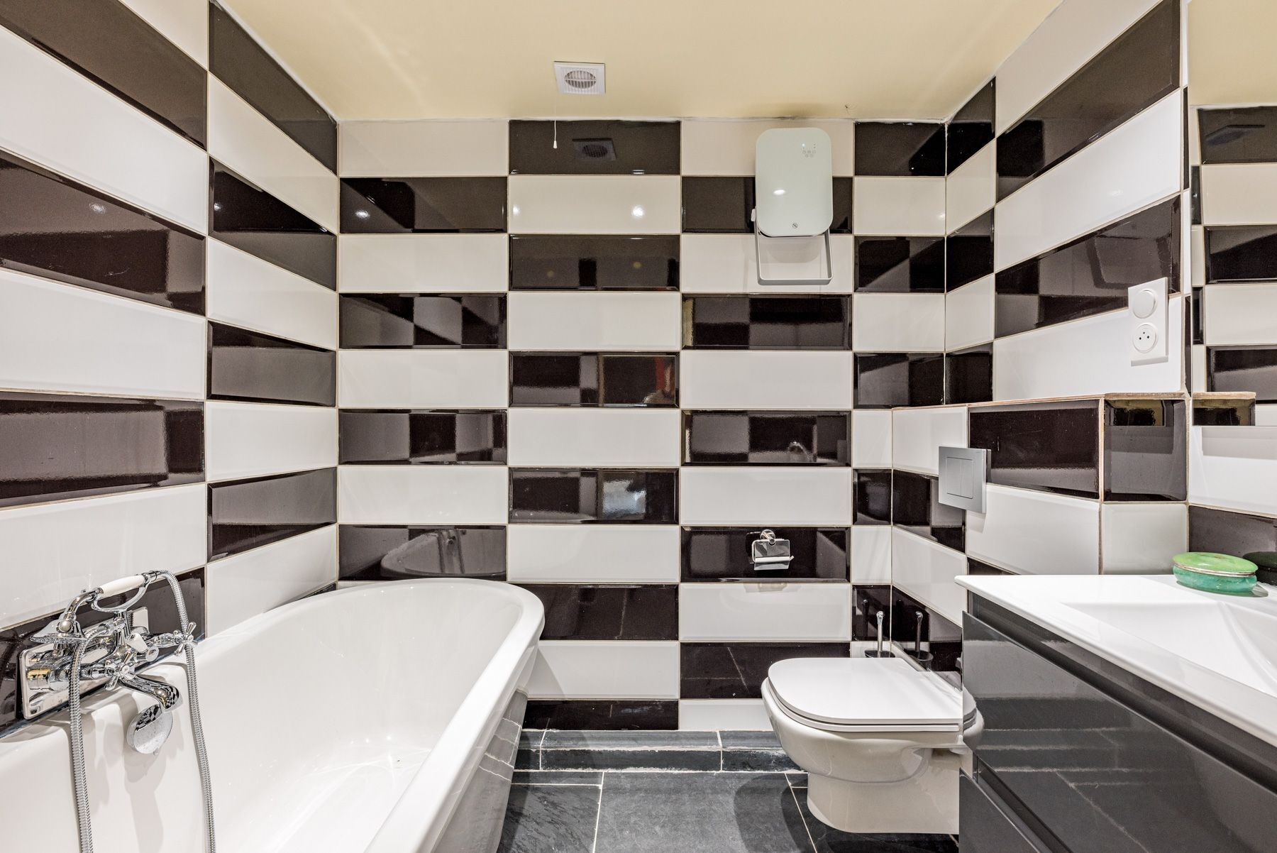 Une salle de bain moderne avec un mur recouvert de carreaux noirs et blancs formant un motif à damier, une baignoire blanche et des toilettes blanches.