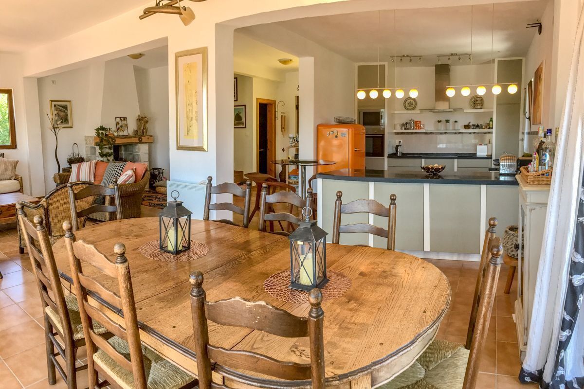 A dining area with a wooden table and chairs, a kitchen in the background, and a small island with a refrigerator.