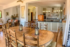 A dining area with a wooden table and chairs, a kitchen in the background, and a small island with a refrigerator.