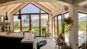 Una cuina àmplia amb una illa gran, zona de menjador amb cadires i vista a un paisatge muntanyós a través de grans finestres.