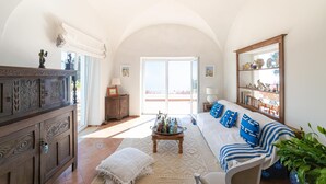 A spacious living room with a high arched ceiling, a large window with a view, a sofa with blue and white striped cushions, and a wooden cabinet with decorative carvings. - Villa Le Tuie by Elite Villas (Capri)
