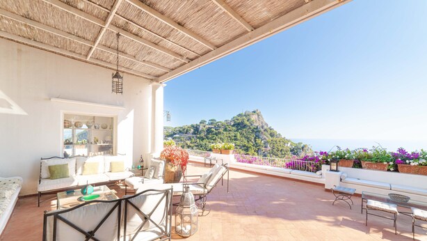 A terrace with outdoor furniture, a view of a mountain, and a white building with a glass door. - Villa Le Tuie by Elite Villas (Capri)