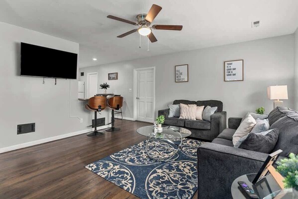 A modern living room with a flat-screen TV, a ceiling fan, a sofa, a coffee table, and a patterned rug.