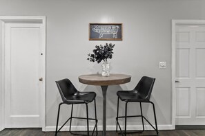 A room with a round table, two black chairs, a vase with black flowers, and a framed sign on the wall.