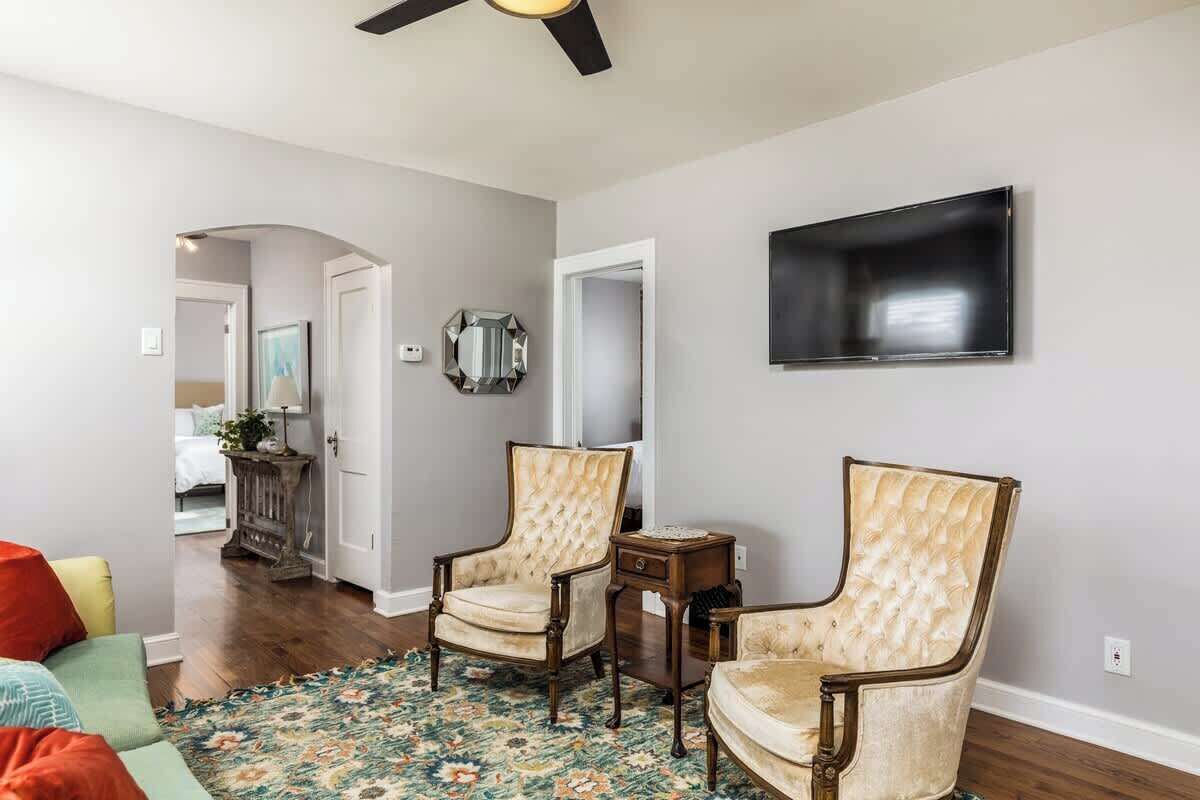 A living room with two upholstered chairs, a floral rug, a flat-screen TV mounted on the wall, and a ceiling fan.