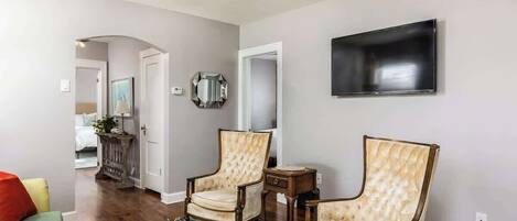 A living room with two upholstered chairs, a floral rug, a flat-screen TV mounted on the wall, and a ceiling fan.