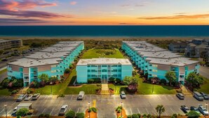 A hotel with a central building flanked by parking lots and a beach view.