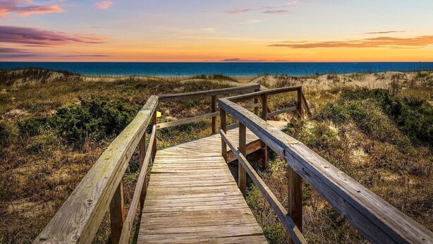A wooden boardwalk leading to a beach with dunes and a sunset in the background.