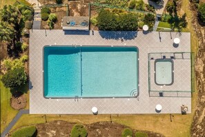 An aerial view of a rectangular swimming pool with a smaller pool adjacent to it, surrounded by a paved deck and greenery.