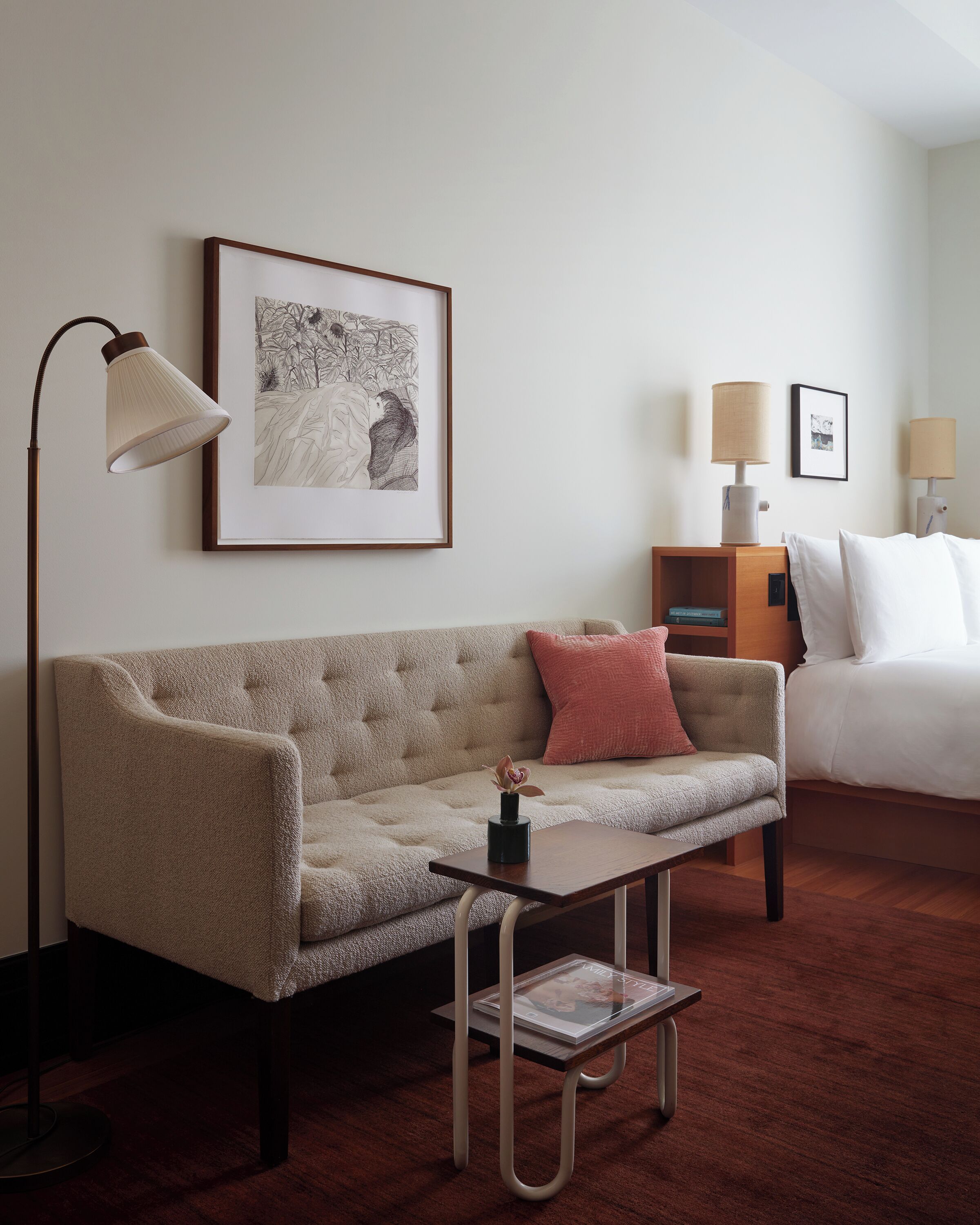 A hotel room with a sofa, a side table, a lamp, a framed picture, and a bed with pillows.