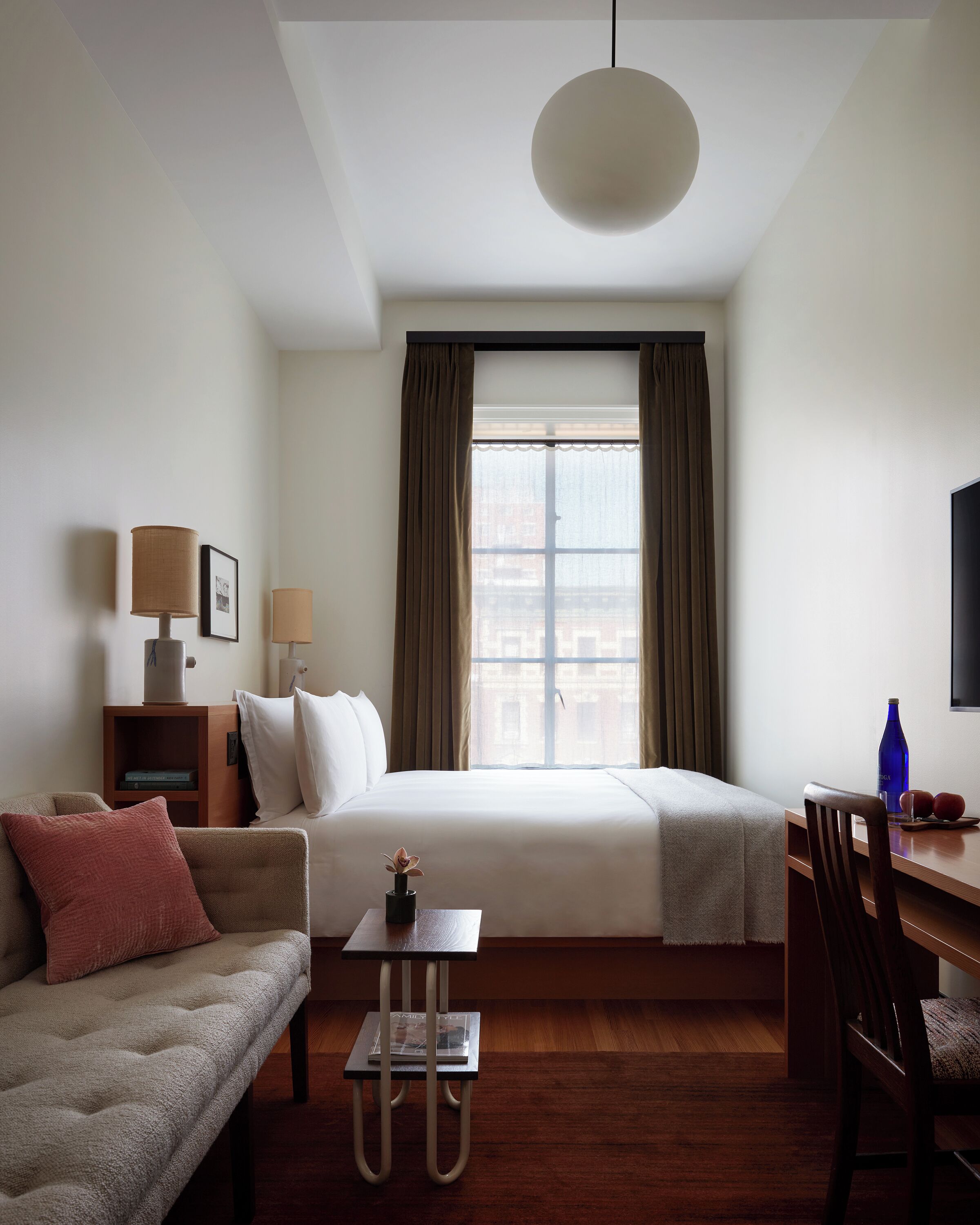 Habitación de hotel con cama, sofá, escritorio, silla, lámpara, un marco de fotos y ventana con cortinas.