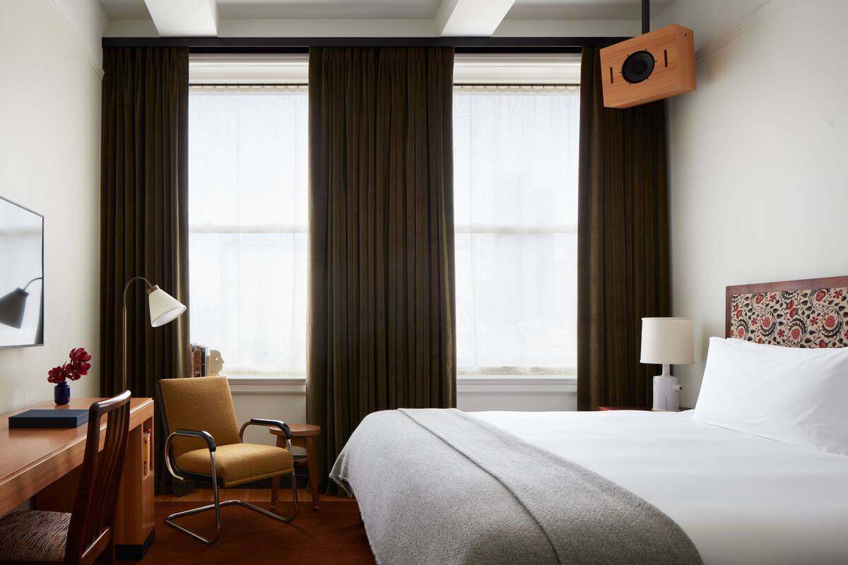 Ein Hotelzimmer mit einem großen Bett, einem Schreibtisch mit Stuhl, einer Schreibtischlampe und einem Wandlautsprecher.