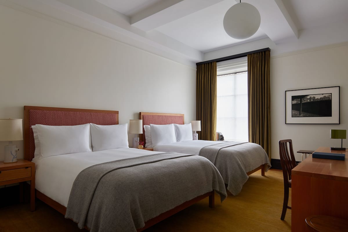 Ein Hotelzimmer mit zwei Betten, einem Schreibtisch, einem Stuhl und einem fensterbepflanzten Fenster.