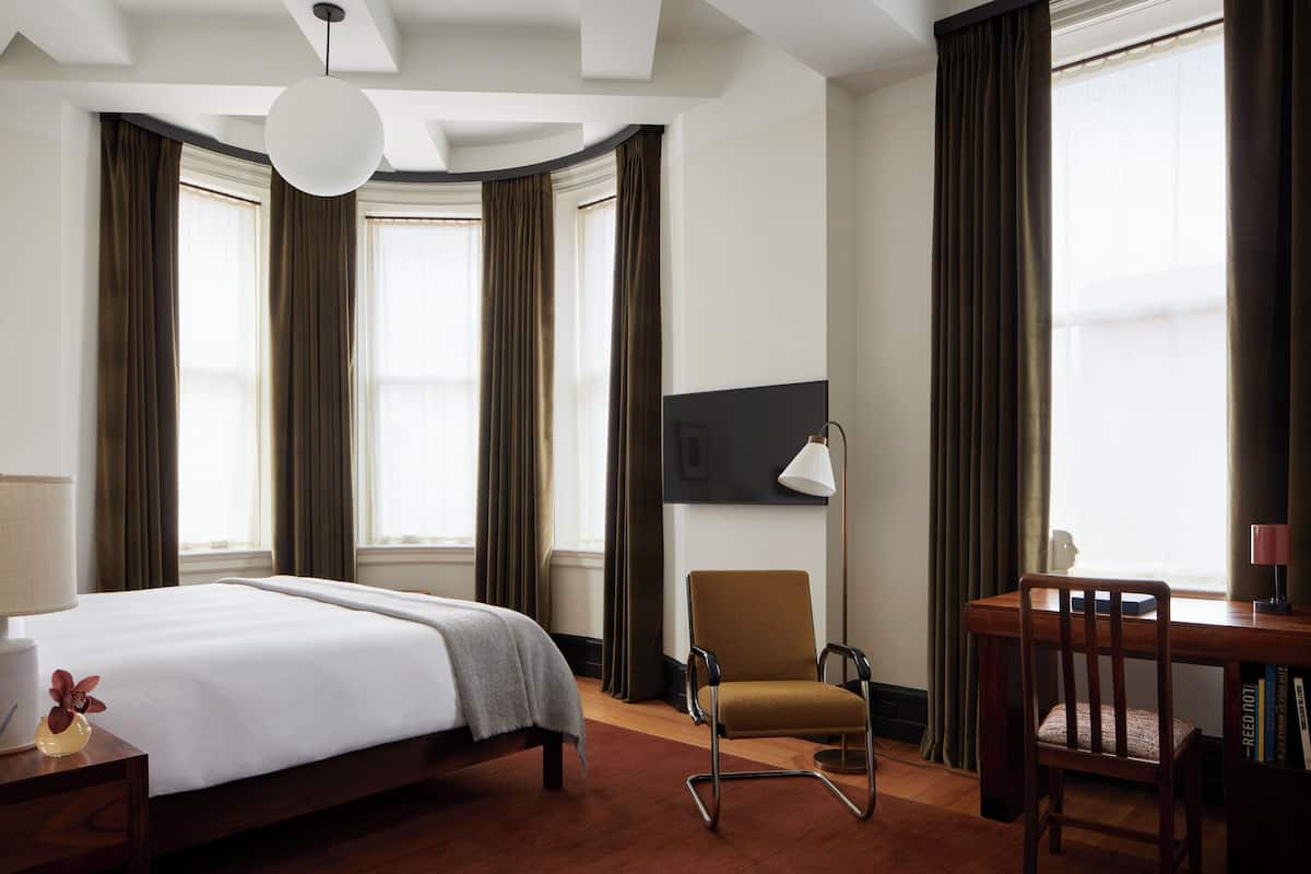 Ein Hotelzimmer mit Bett, Schreibtisch, Stuhl, Lampe und einem großen Fenster mit Vorhängen.